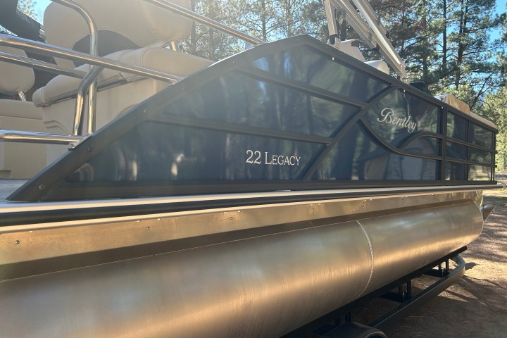 Side view of a Bentley 22 Legacy pontoon boat on a trailer in a wooded area.